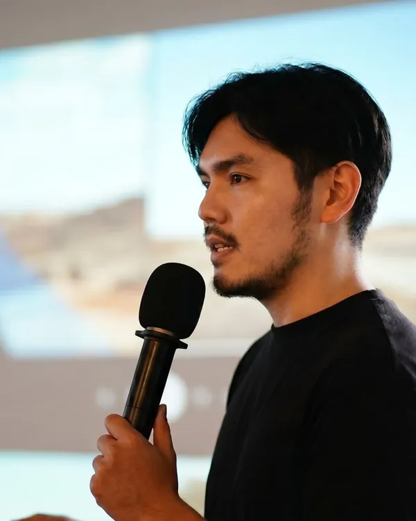 葉議方穿黑色上衣，手持麥克風側身演講，背景為大型投影畫面