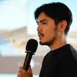 葉議方穿黑色上衣，手持麥克風側身演講，背景為大型投影畫面